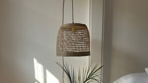 Ett träbord som står vid sängen, med en flätad lampa och en vas med en växt i.
