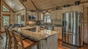 A kitchen with wooden cabinets, a stainless steel refrigerator, granite countertops, and a large island with bar stools. - Raven’s Retreat: Sauna, Putting Green & Arcade! (Blue Ridge)