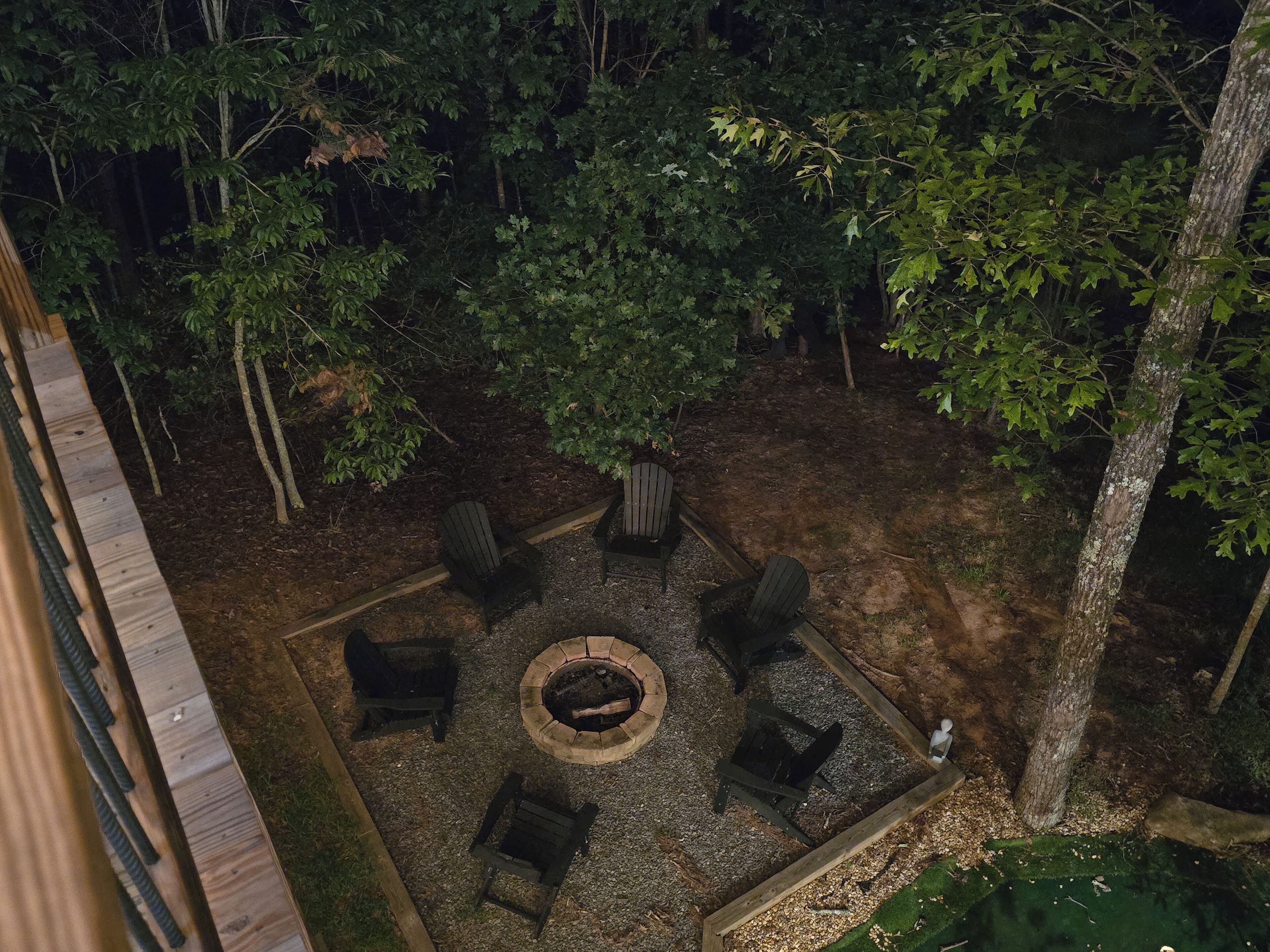 An outdoor seating area with a fire pit surrounded by chairs and tables, set in a wooded environment.