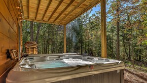 A hot tub with a wooden exterior, surrounded by a wooden deck and a forest view. - Raven’s Retreat: Sauna, Putting Green & Arcade! (Blue Ridge)