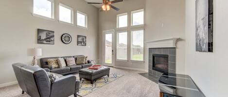 A spacious living room with a fireplace, a ceiling fan, and large windows.