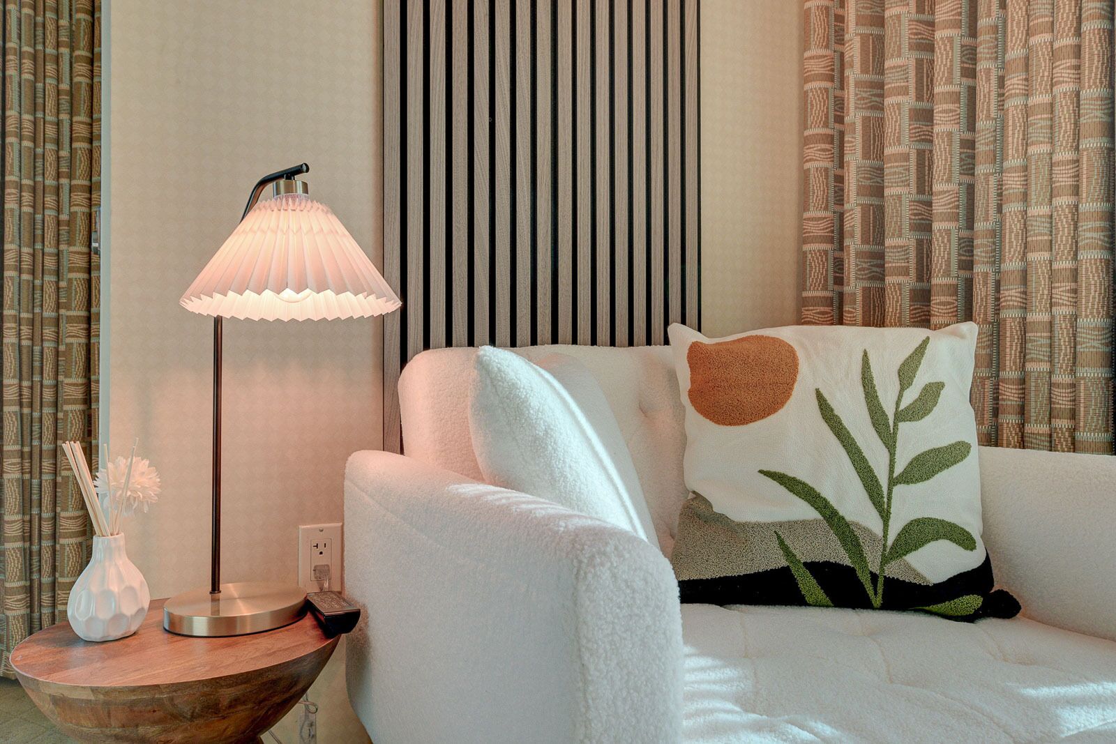 Een moderne kamer met een witte bank, een houten bijzettafeltje met een lamp en kamergordijnen met een patroon.