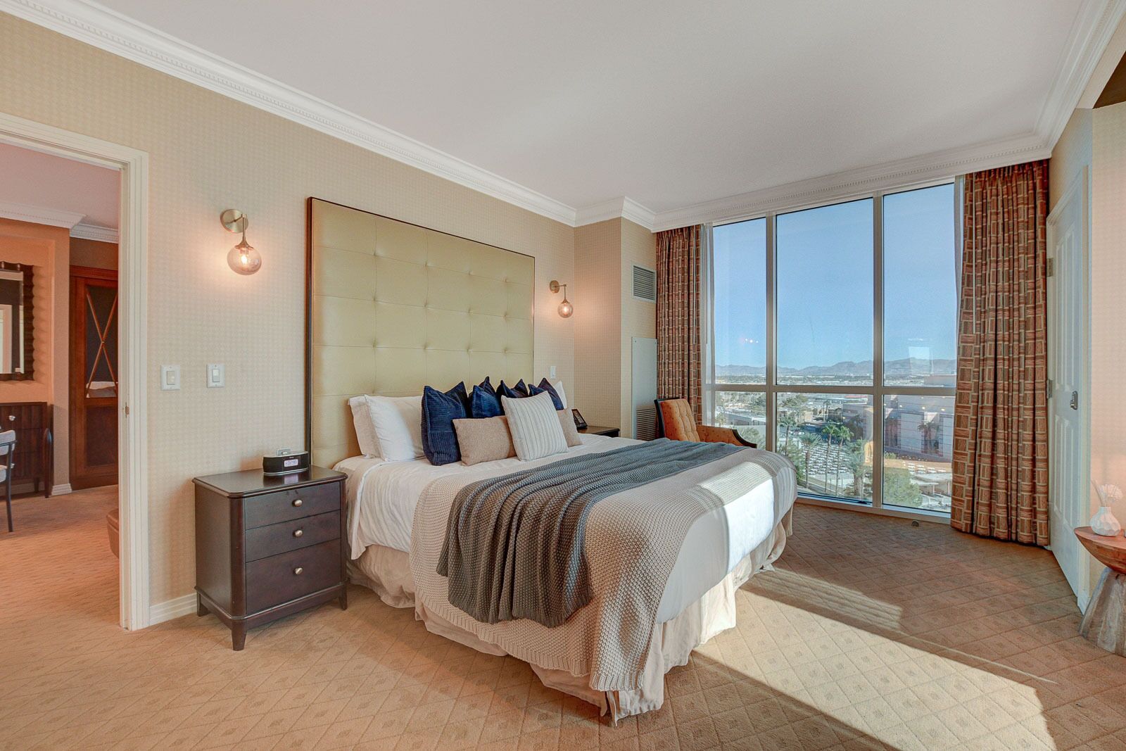 A spacious bedroom with a large bed, a nightstand, a dresser, and a view of the city through large windows.