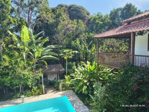 A pool surrounded by lush greenery, a thatched-roof structure, and a building with a tiled roof. - Casa Recanto da Mata, Localizada em Trancoso, Condomínio Coqueiral km 6 (Trancoso)