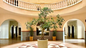Een ruime lobby met een vloer met geometrisch patroon, een grote potplant in het midden en een gebogen balkon met balustrades.