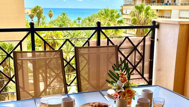 Een balkon met een tafel, ontbijtartikelen, een bloevas en uitzicht op zee en palmbomen.