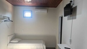 A small, single-bed room with a wooden ceiling, a window, an air conditioning unit, a TV mounted on the wall, and a desk with a chair. - Pousada Paraiso (Wanderley)