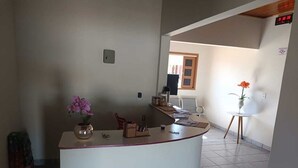 A reception area with a counter, a potted plant, a small table with a vase, and a window with curtains. - Pousada Paraiso (Wanderley)