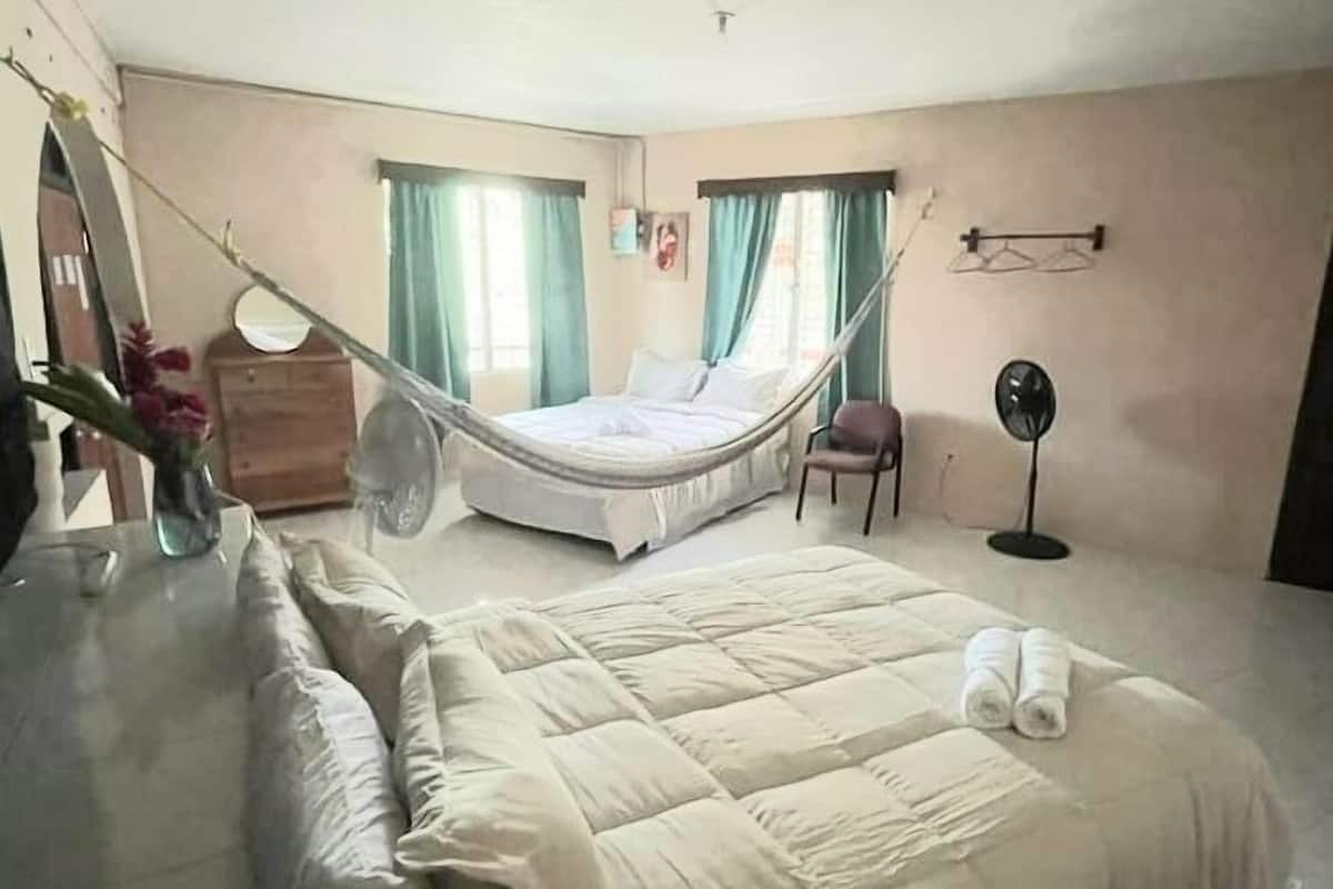 A bedroom with a large bed, a bedside table, a chair, a standing fan, and a window with curtains.