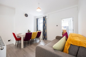 A modern living room with a sofa, dining table, and chairs. There is a window with curtains, a clock on the wall, and a door leading to another room. - Manor Terrace Retreat Accrington (Accrington)