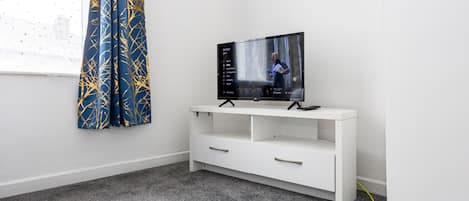 A room with a white entertainment unit, a flat-screen TV, a grey carpet, and a window with a curtain.