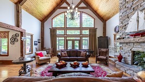 A spacious living room with a high wooden ceiling, large windows, a fireplace, and a cozy seating area. - 8 bed 5 bath cottage on Lake Muskoka, 9 acres! (Torrance)