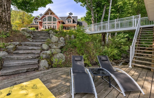 A wooden deck with lounge chairs, a yellow mat, and a stone staircase leading to a house with a white railing.