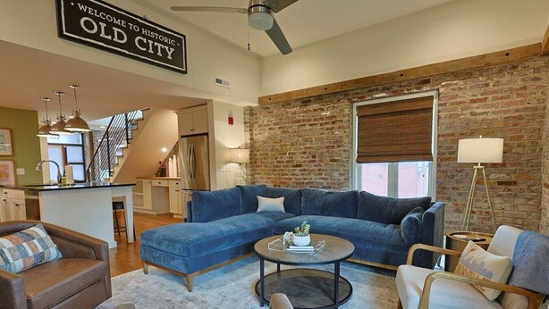 A living room with a blue sectional sofa, a round coffee table, and a brown leather armchair. There is a brick wall and a ceiling fan.