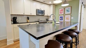 A kitchen with white cabinets, a black countertop, a stainless steel microwave, and a sink. There are three stools at the island.