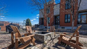 An outdoor seating area with Adirondack chairs and a fire pit, surrounded by a brick patio and a building with large windows.