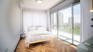 Un dormitorio moderno con una cama grande, una mesita de noche y vistas a un paisaje urbano a través de ventanales del suelo al techo.