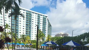 A resort pool area with lounge chairs, umbrellas, and a large hotel building in the background.