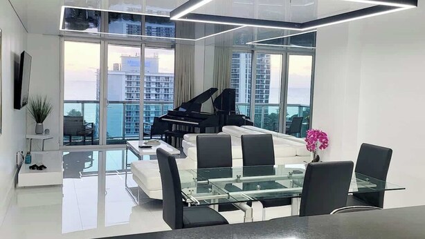 A modern living room with a glass dining table, black chairs, a piano, and a large window offering a city view.