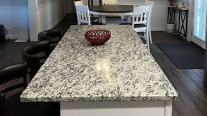 Une cuisine dotée d’un plan de travail en granit, de meubles blancs, d’un ventilateur de plafond et d’un téléviseur fixé au mur.