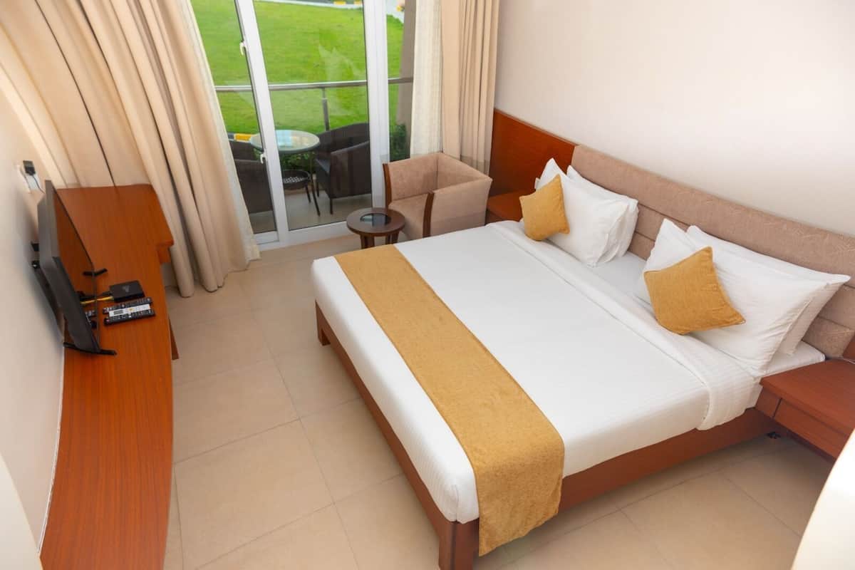 A hotel room with a large bed, a desk with a computer, a small table, and a view of a green lawn through a glass door.