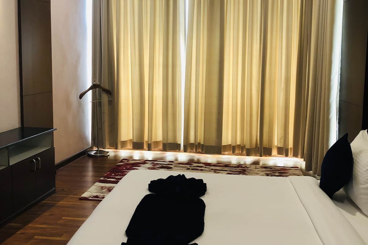 A hotel room with a neatly made bed, a dark wooden wardrobe, a floor lamp, and a large window with curtains.