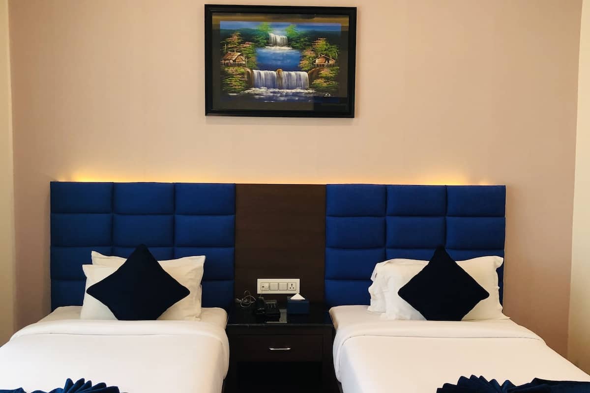 A hotel room with two beds, a blue headboard, a painting of a waterfall, and a nightstand with a phone and a tissue box.