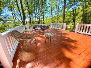 Una terraza de madera con muebles de mimbre y una mesa de vidrio, rodeada de barandillas blancas y árboles.