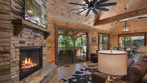 A cozy cabin interior with a stone fireplace, wooden ceiling, and a dining area with outdoor views. - Stunning Mountain View With Hot tub & Game Room. Indoor/Outdoor Fireplace (Ellijay)
