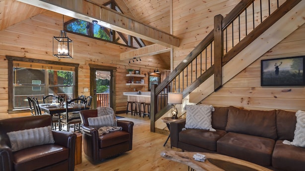 A spacious log cabin interior with a dining area, a staircase, and leather furniture. - Stunning Mountain View With Hot tub & Game Room. Indoor/Outdoor Fireplace (Ellijay)