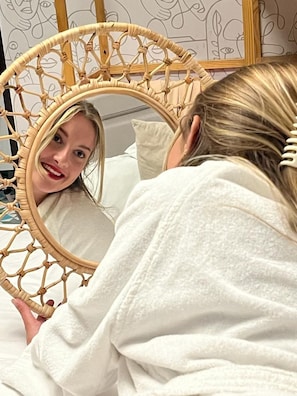 A person in a white robe sitting in front of a mirror with a woven frame.