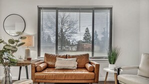 A living room with a brown leather sofa, a round mirror, a lamp, and a window with blinds.