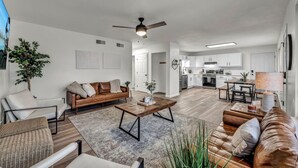 A spacious living room with a brown leather sofa, a wooden coffee table, and a ceiling fan. A kitchen with white cabinets and a dining area with a table and chairs is visible in the background.