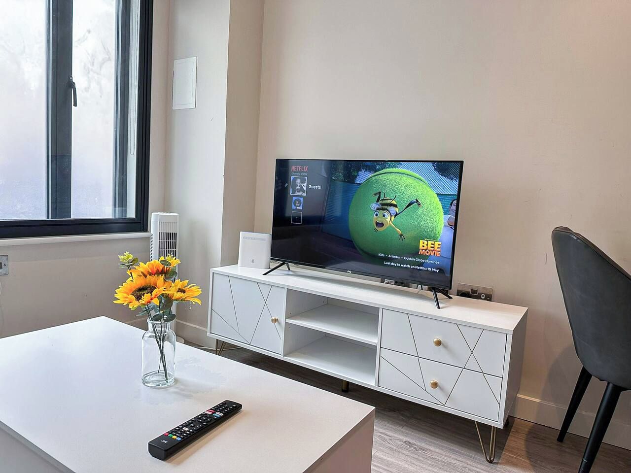 Een moderne woonkamer met een flatscreen televisie op een wit tv-meubel, een vaas met zonnebloemen op een witte tafel, en een afstandsbediening.