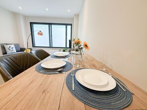 Et spisebord dækket med tallerkener, bestik og glas, med udsigt til et vindue og en sofa i baggrunden.
