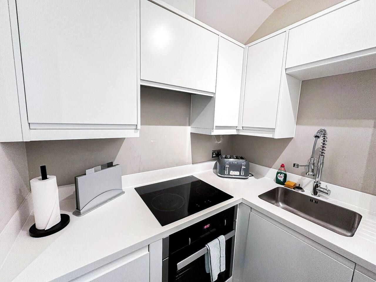 A modern kitchen with white cabinetry, a black induction cooktop, a built-in oven, a stainless steel sink, and a toaster.