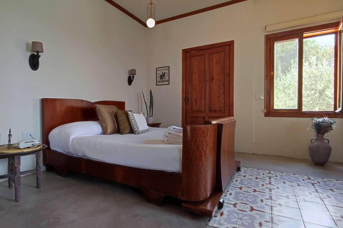 A bedroom with a wooden bed, a small bedside table, a nightstand, a window with a view of trees, and a vase with flowers.