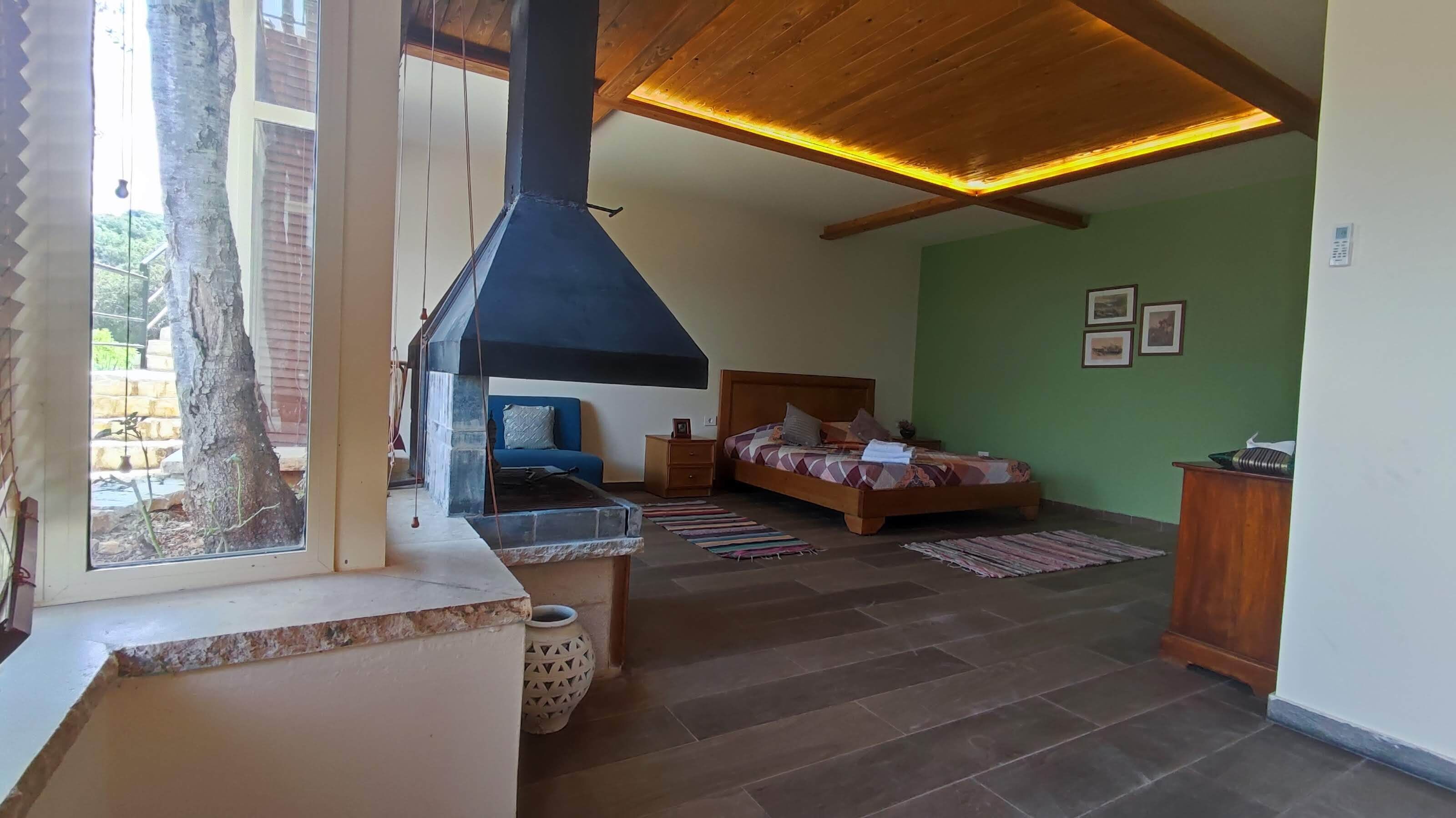 A bedroom with a bed, a blue sofa, a wooden cabinet, and a large window overlooking a stone wall.