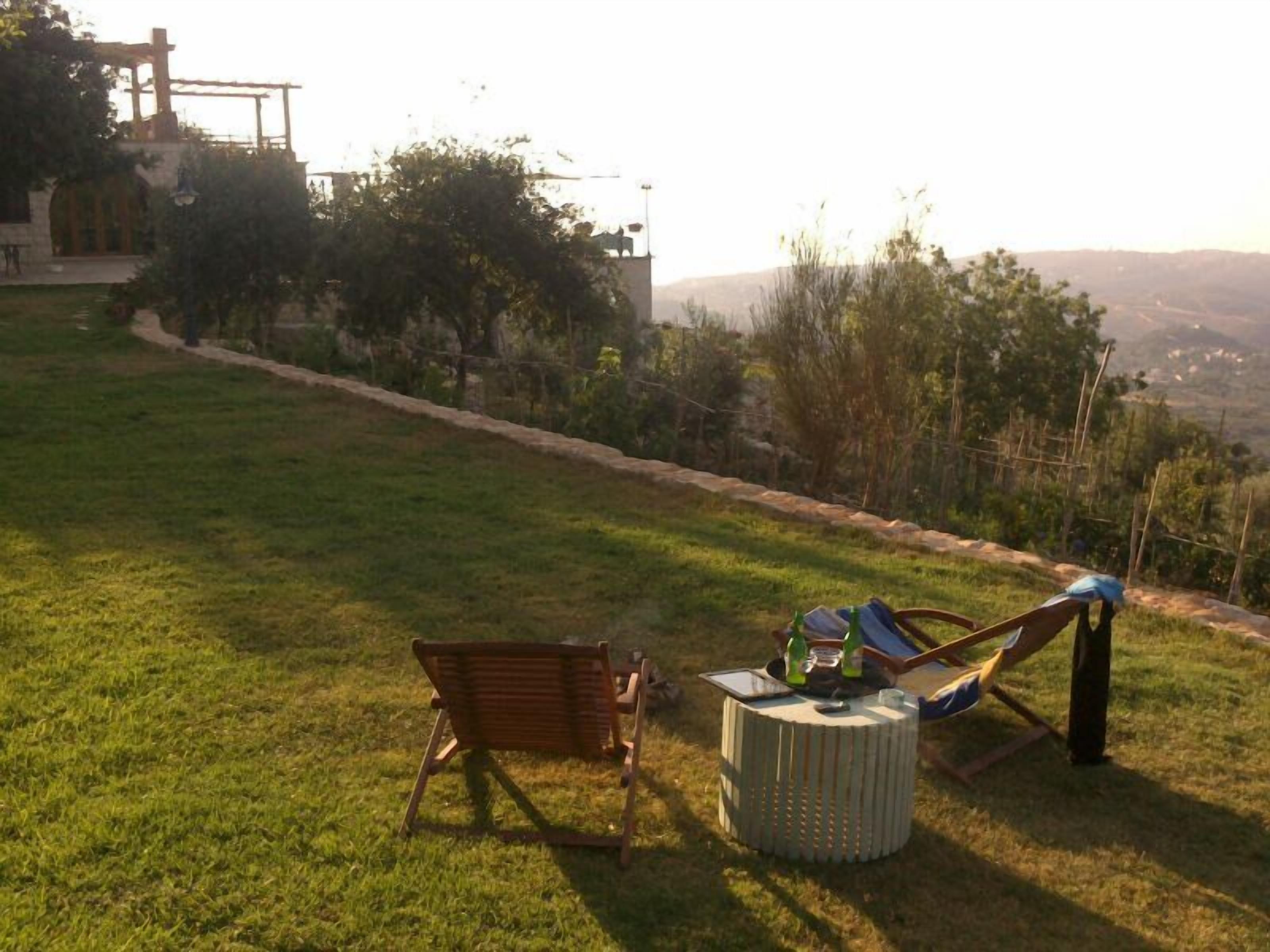 Zona relax all'aperto con una sedia di legno e una poltrona prendisole, un tavolino con una bevanda e vista su un paesaggio collinare.