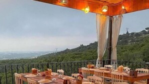 Un área de comedor al aire libre con mesas y sillas de madera, con vistas panorámicas a colinas y un paisaje urbano distante.