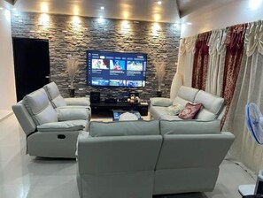 A modern living room with a stone wall, two grey sofas, a flat-screen TV, and a fan. - Bella villa in Djidjole with Safety Keeper (Accra)