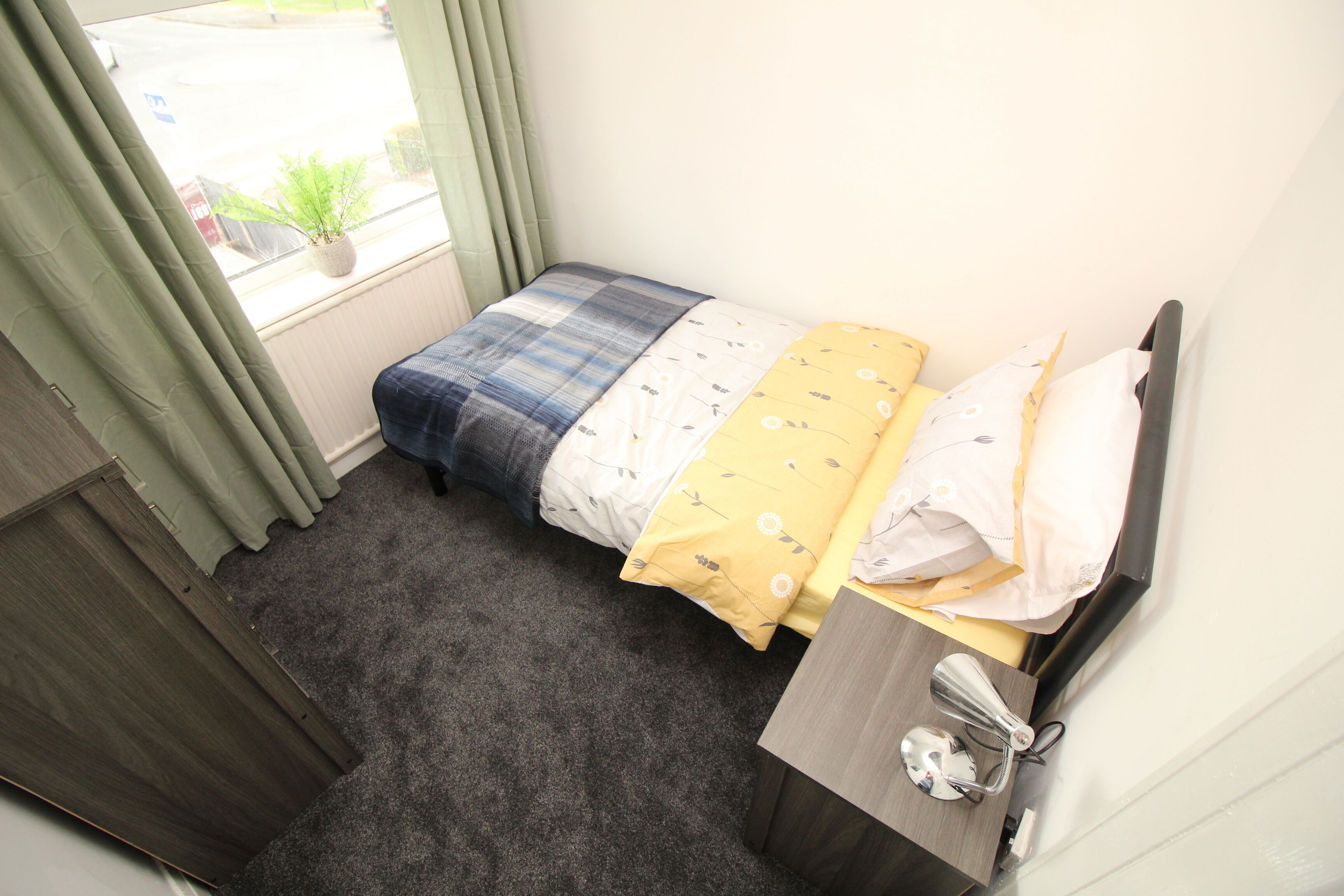 Ein kleines, ordentlich eingerichtetes Schlafzimmer mit Bett, Nachttisch, Spiegel und einem Fenster mit Vorhängen.