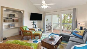 A spacious living room with a ceiling fan, a flat-screen TV, wicker furniture, a sofa, and a view through large sliding glass doors to an outdoor deck. - Perch in Paradise on St Simons Island (St. Simons Island)