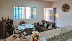 A living room with a sofa, a dining table with chairs, a coffee table with fruit, and a bookshelf. - Casa Aconchegante em Santa Cruz Cabrália -Ba (Santa Cruz Cabrália)