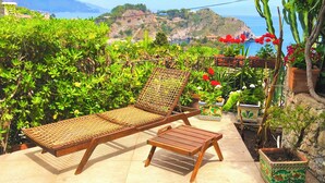 A poolside area with two wooden lounge chairs, a small wooden table, and potted plants.