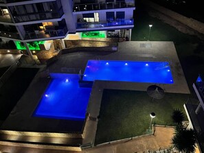 Un edificio de varios niveles con área de piscina, que incluye una piscina grande y una más pequeña, rodeadas por un jardín ajardinado.