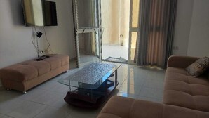 Uma sala de estar com sofá, puf e mesa de centro. Há uma televisão na parede e uma porta de vidro deslizante que dá para o exterior.