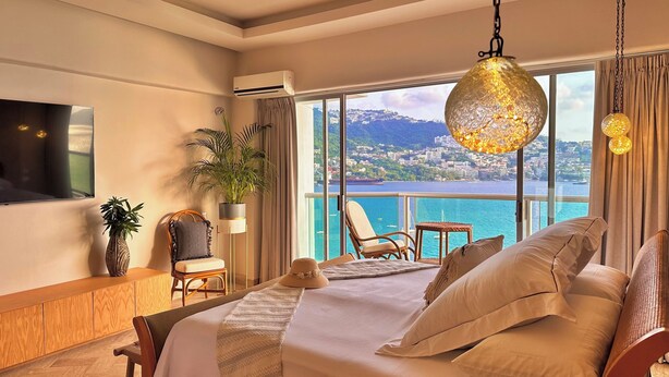 A hotel room with a large bed, a TV mounted on the wall, a potted plant, a chair, and a view of the sea.