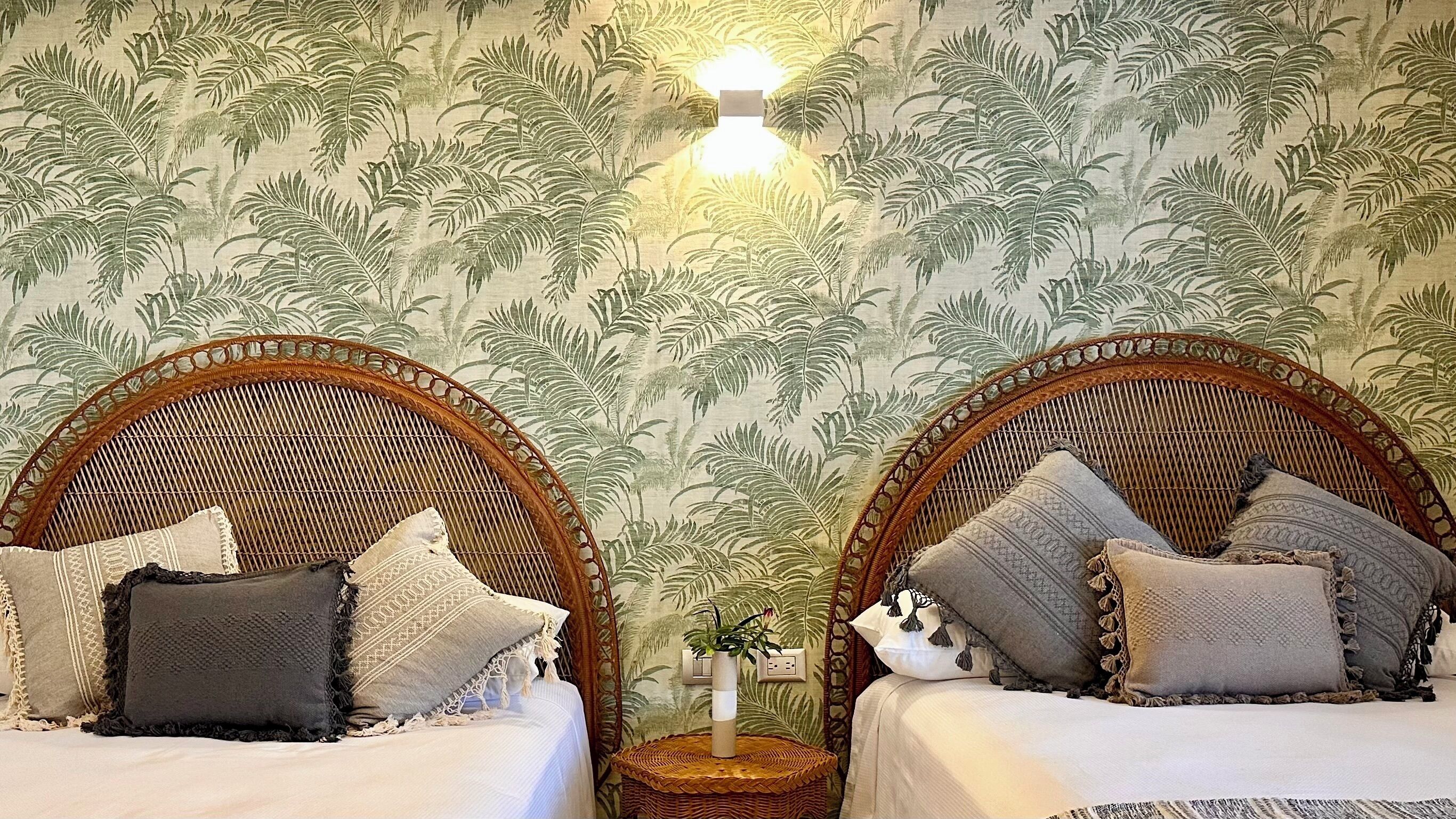 Une chambre avec deux lits en osier, chacun recouvert d’une courtepointe blanche et agrémenté de plusieurs coussins. Une petite table en osier, sur laquelle est posé un vase de fleurs, se trouve entre les deux lits.