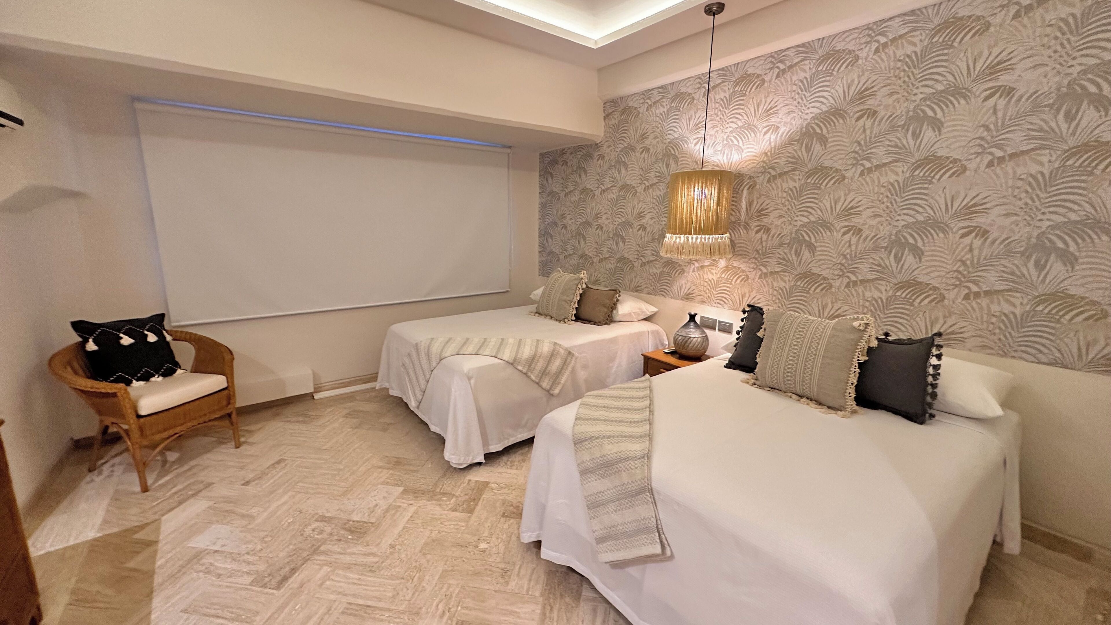 Quarto com duas camas, uma cadeira de madeira, uma janela grande e parede com padrão.